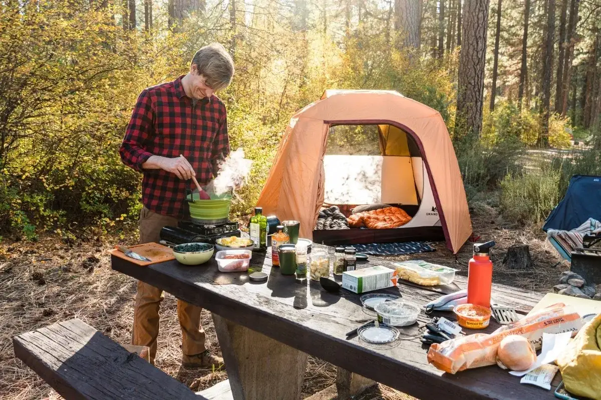 Man cooks a delicious, cozy camping food meal outdoors next to a tent, with ingredients spread across a picnic table.