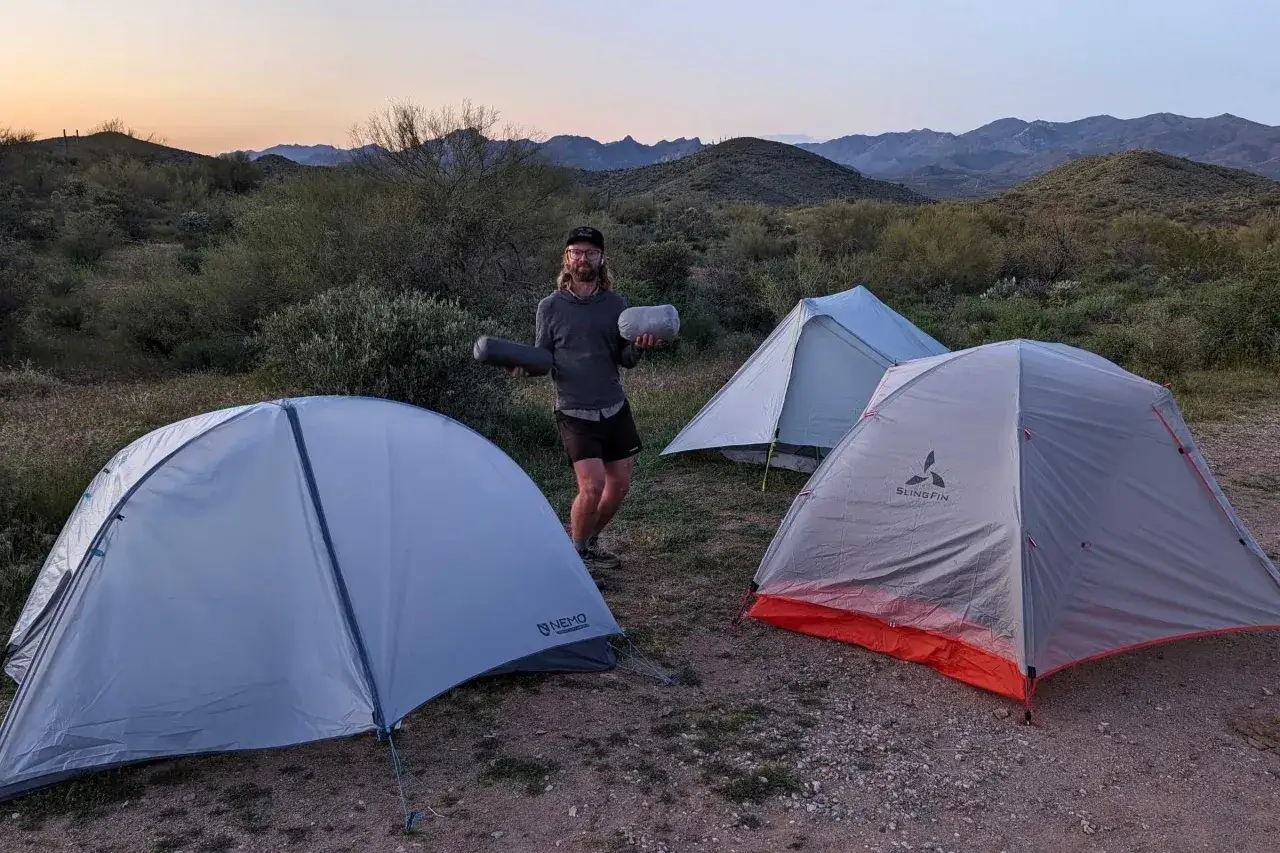 A man holds sleeping bags, ready for a gossamer gear the two review. Three tents are set up in a desert landscape at dusk.