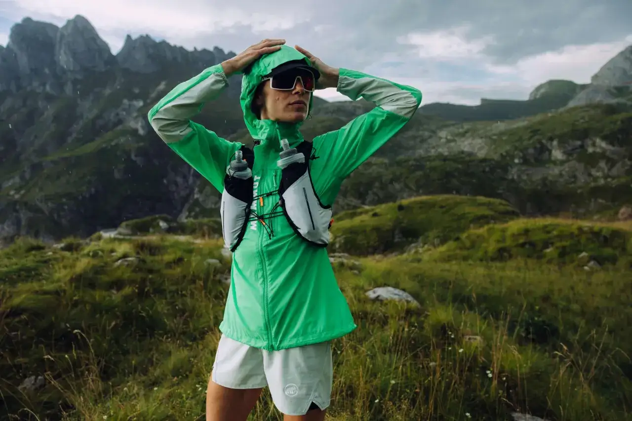 A hiker in a green jacket and shorts, wearing a hydration vest with water bottles, poses in a mountainous landscape. This image reminds us how to stay hydrated while hiking.