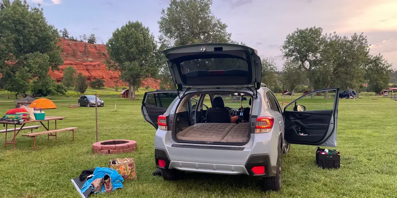 Car camping for beginners: an air mattress fills the back of a car, ready for a night under the stars. Gear is laid out on the grass near a picnic table and fire pit.