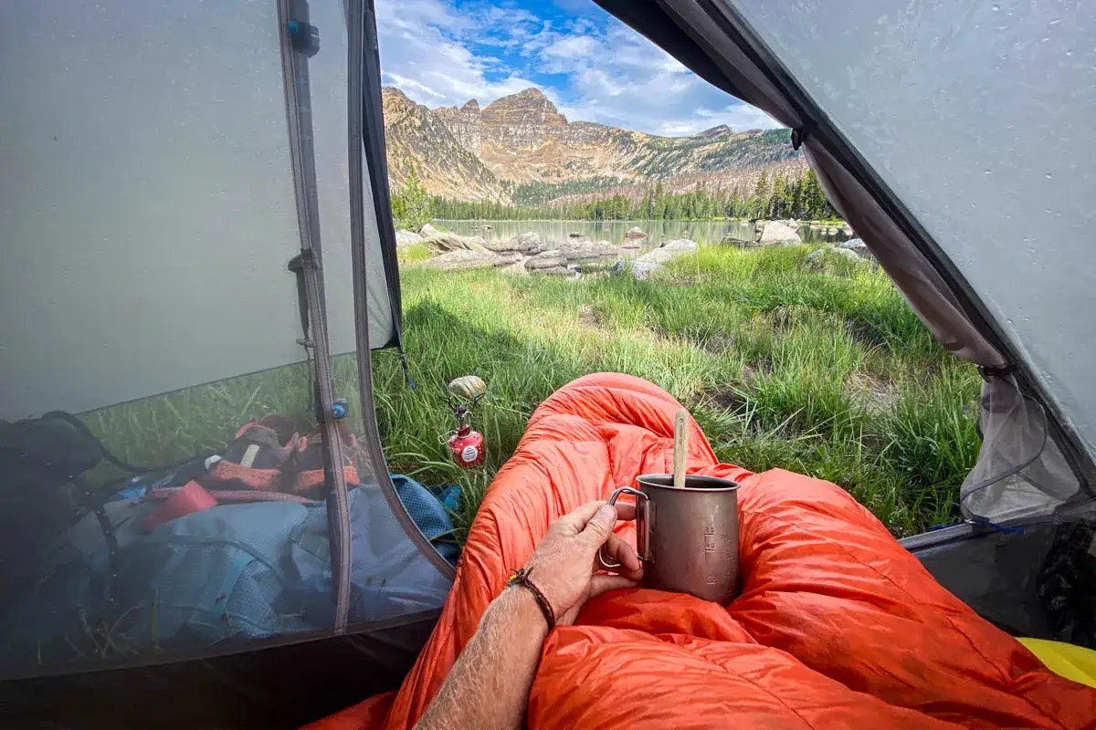 Cozy in a Z-pack 20-degree sleeping bag, enjoying a warm drink with a stunning mountain lake view from the tent.
