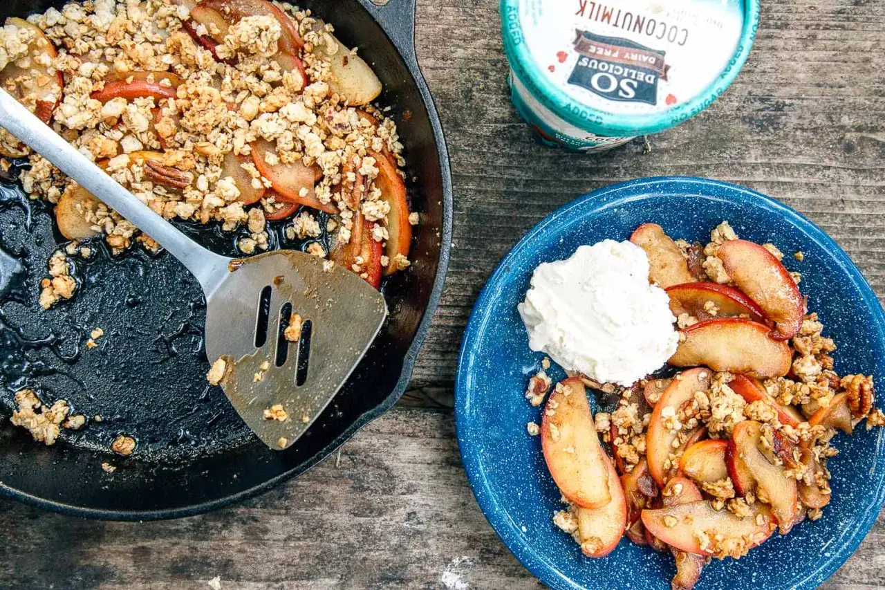 A warm campfire apple crisp, served in a cast iron skillet and a blue enamel plate, topped with a dollop of whipped cream.