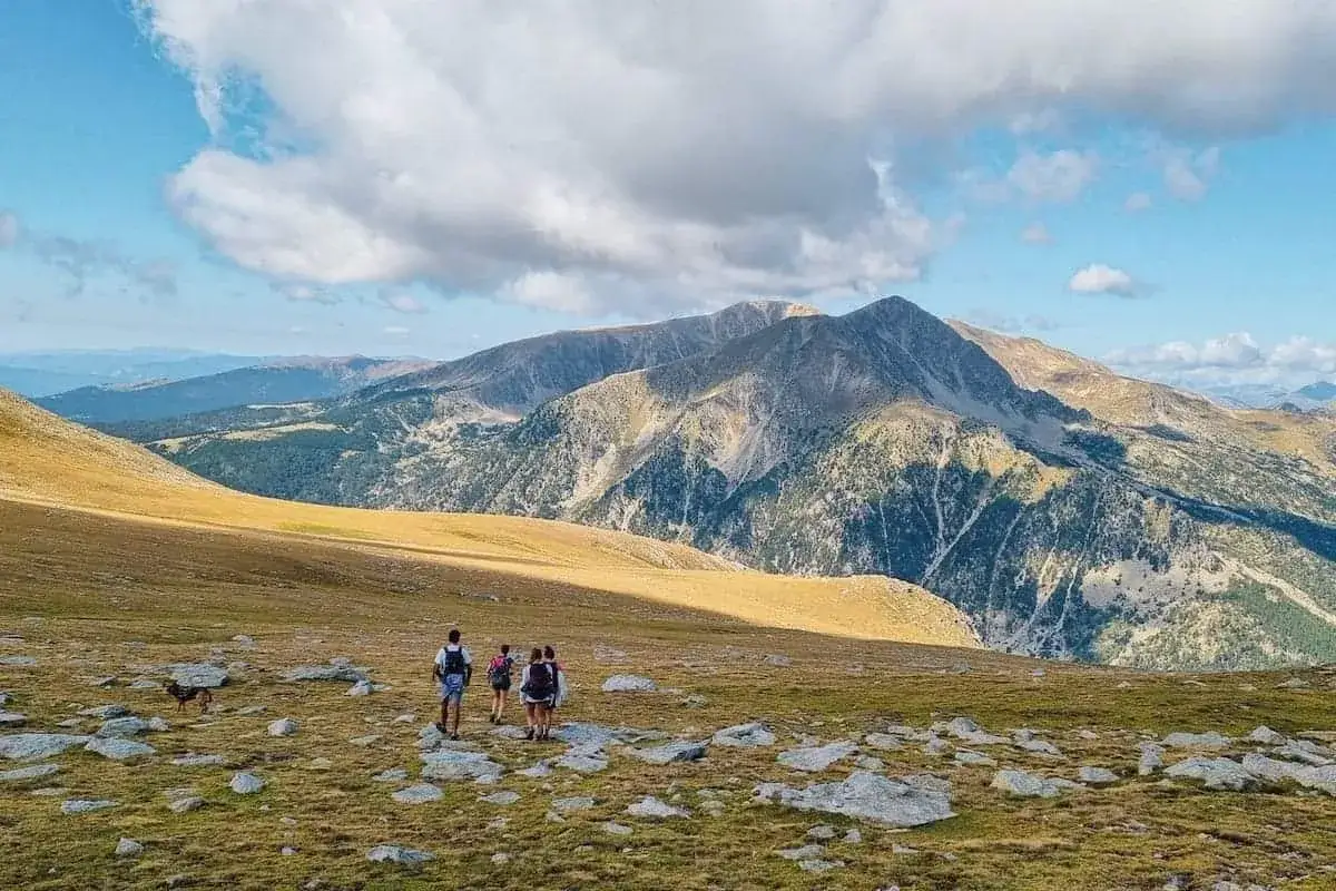 Hikers with backpacks traverse a rocky, grassy meadow with a dog, on a pyrenees hut to hut hiking adventure. Majestic mountains rise in the background under a cloudy sky.