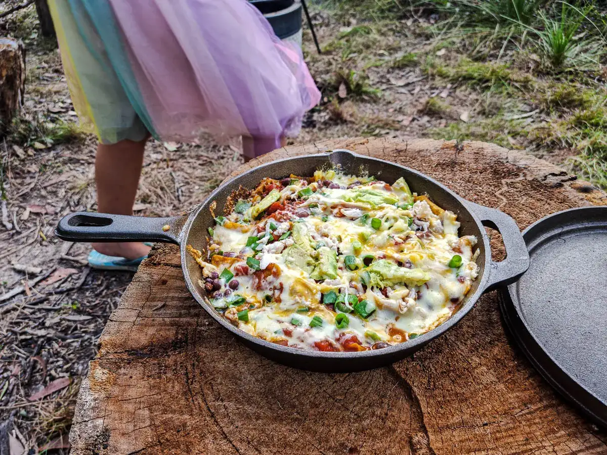Delicious camping nachos, bubbling with cheese and topped with avocado and green onions, are served in a cast-iron skillet on a tree stump.