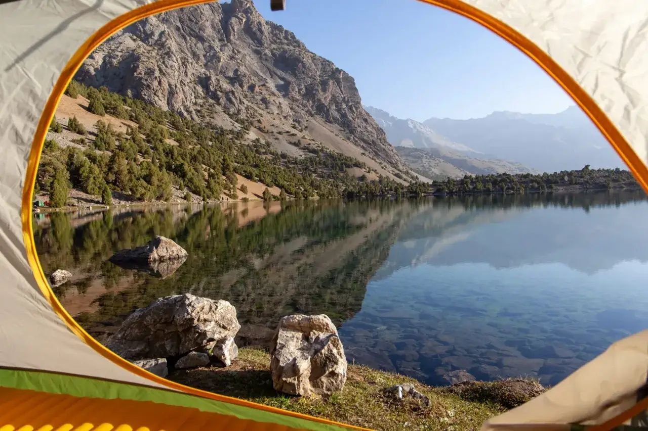 View from a tent at a serene mountain lake, perfect for backpacking hiking. The clear water reflects the rugged slopes and sparse trees.