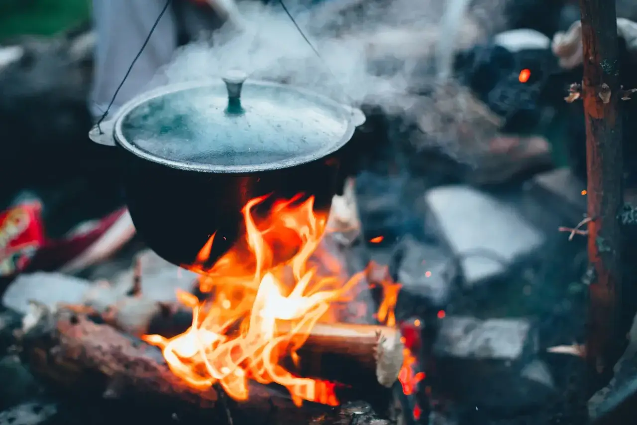 A black pot hangs over a campfire, steam rising, promising cozy camping food. Flames lick the pot, creating a warm, inviting scene.