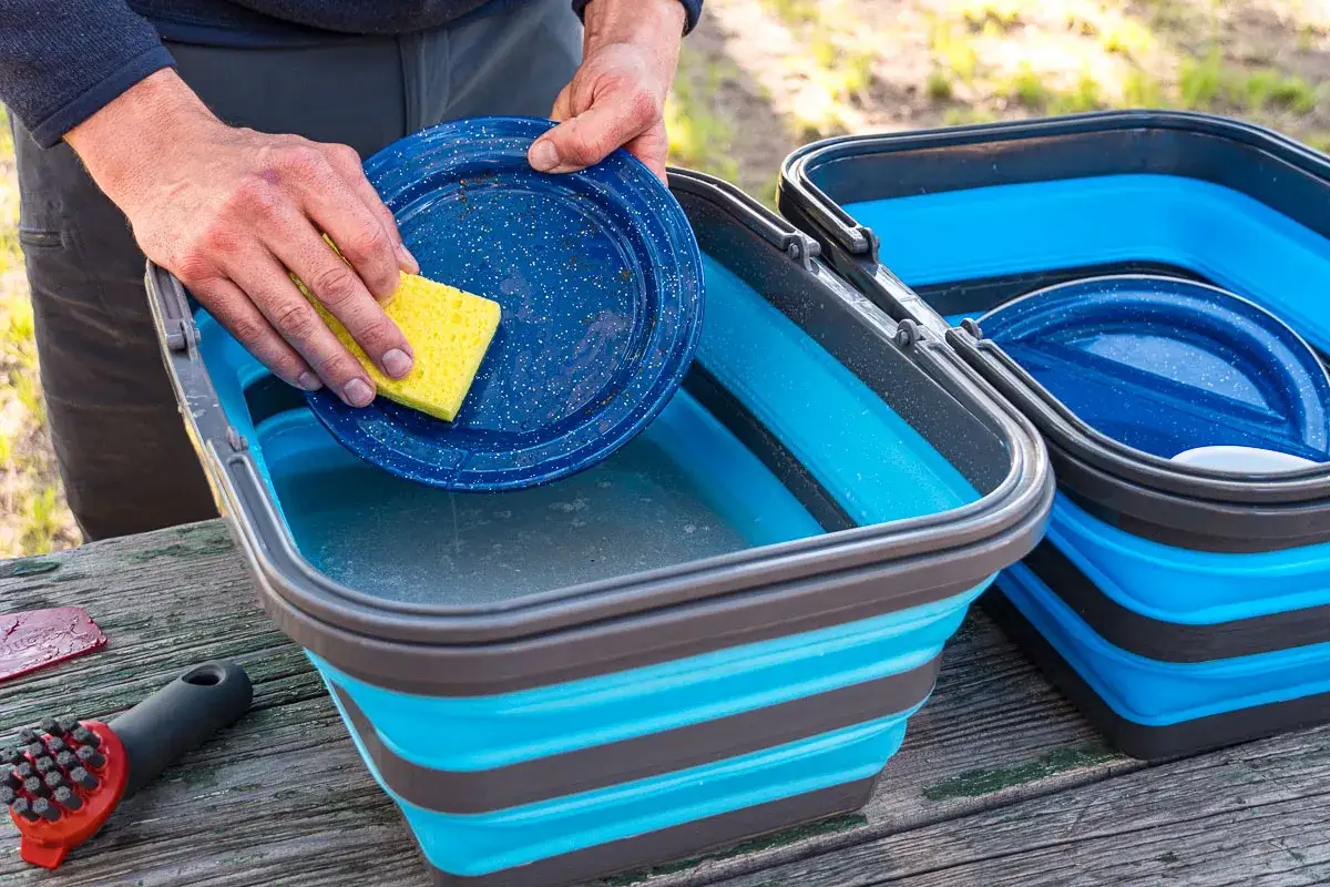 Hands washing a blue speckled plate with a yellow sponge in a collapsible blue and gray camping wash basin.