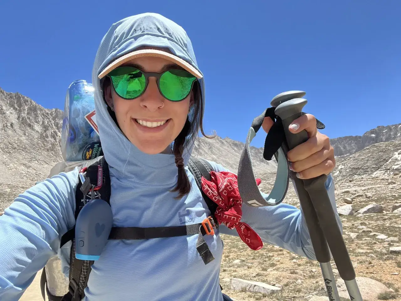 A hiker smiles, wearing a light blue hooded shirt, sunglasses, and a bandana, ready for a hike in hot weather with trekking poles and a backpack.