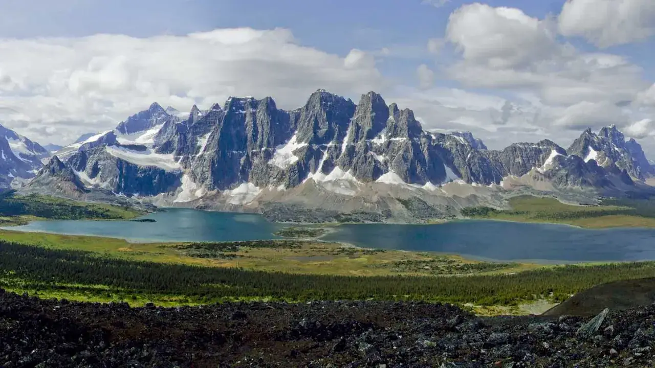A breathtaking panorama of the Tonquin Valley hike, featuring jagged, snow-capped mountains reflecting in a serene blue lake, surrounded by lush green meadows and dark, rocky foreground.