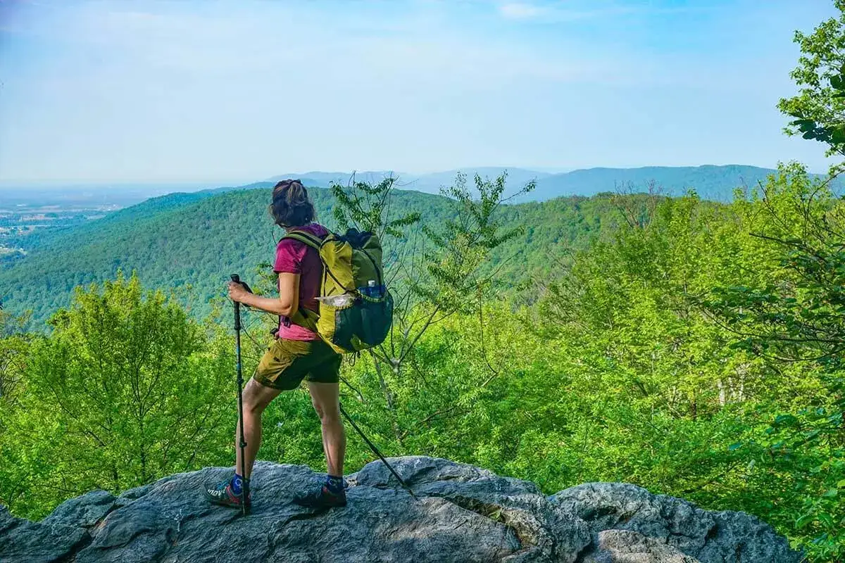 A hiker with a backpack and poles stands on a rocky outcrop, enjoying a scenic view. This is excellent training for hiking.