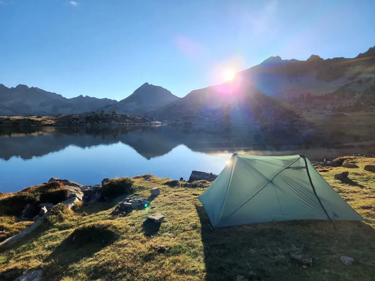 A green tent sits by a calm lake, reflecting mountains under a bright sunrise. A perfect spot for an hrp trail adventure.