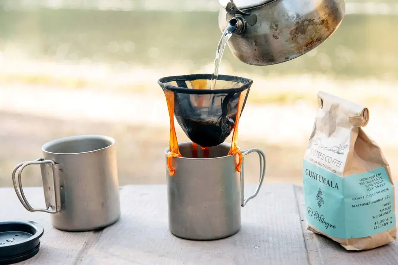 Making coffee while camping: hot water is poured from a kettle into a pour-over filter set over a mug, with a bag of Guatemala coffee nearby.