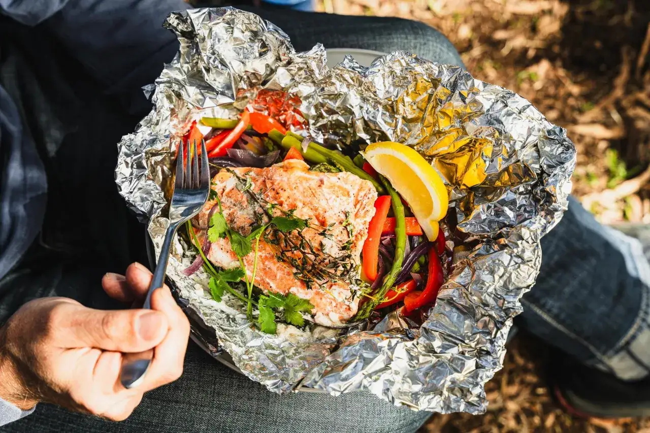 A fork is about to dig into a foil-wrapped dinner of salmon and vegetables. Curious how long to cook tin foil dinners in fire? This looks delicious!