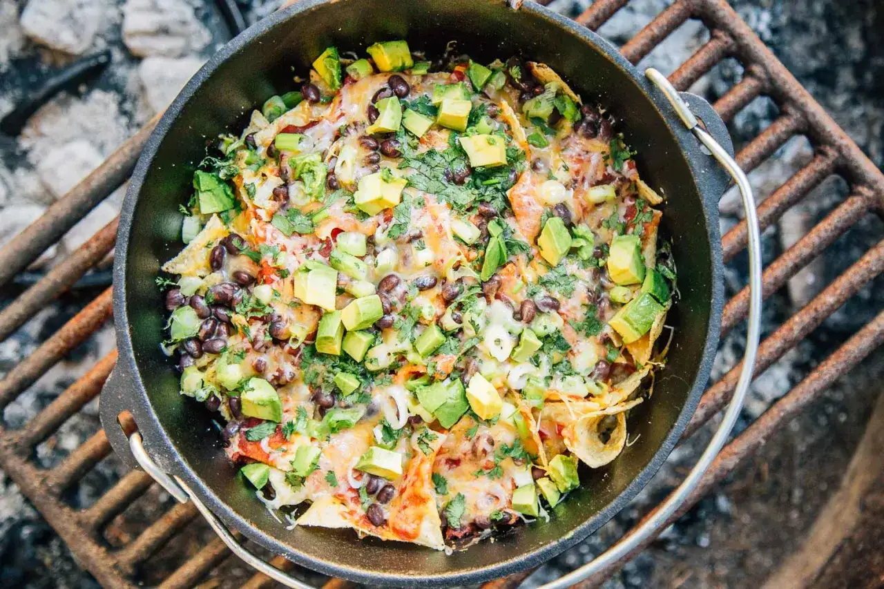 Melted cheese, black beans, and avocado top crispy tortilla chips in a cast-iron skillet, a perfect example of easy camping meals cooked over a fire.