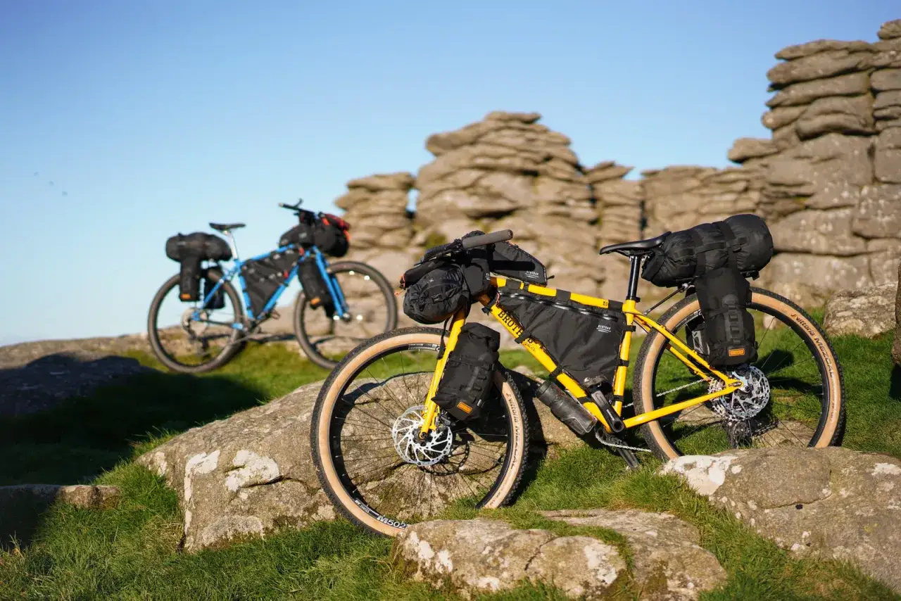 Two fully loaded bikepacking setup bikes rest on a rocky, grassy hillside under a clear blue sky.