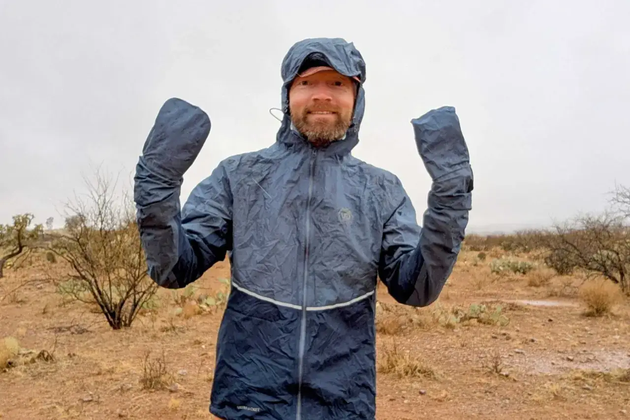 A man in a desert landscape smiles, holding up his arms covered by ultralight waterproof jackets.
