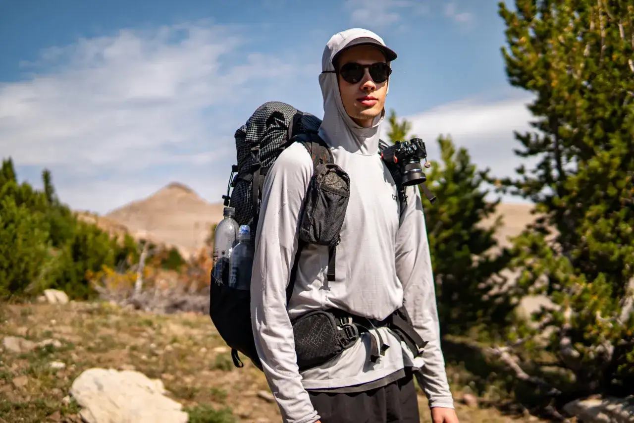 A young man with sunglasses and a backpack is hiking in hot weather, with a camera attached to his pack.