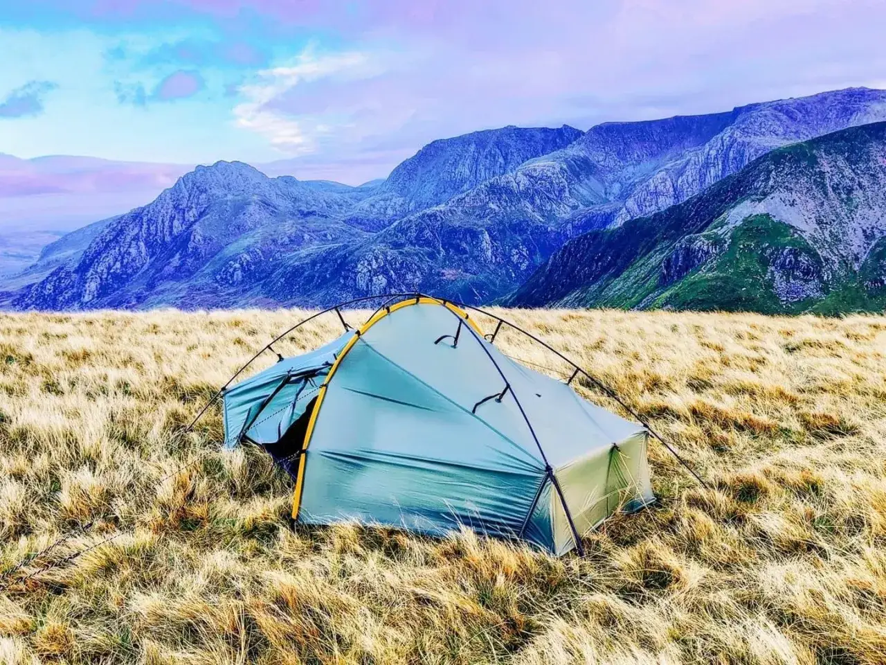 A tent pitched in a field of dry grass, with a majestic mountain range in the background. This is how to camp in the wild.