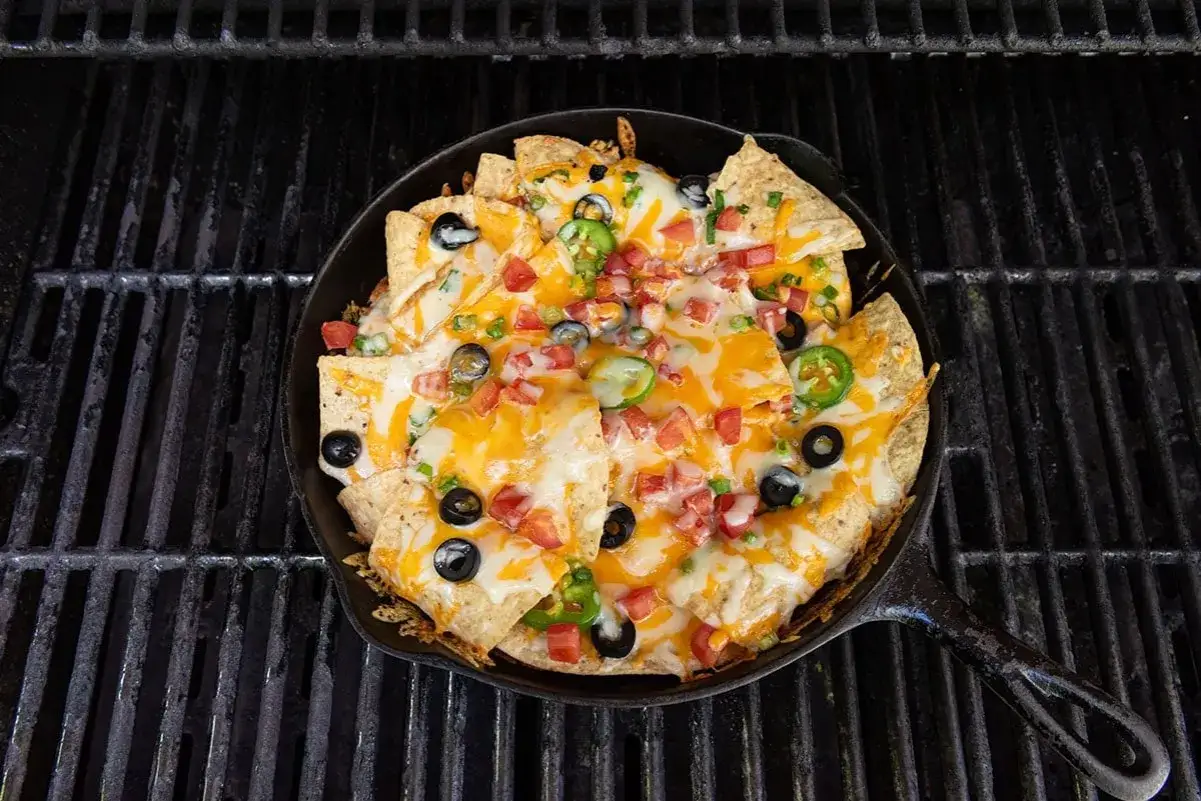 Camp taco perfection: a cast iron skillet overflowing with cheesy nachos, jalape&ntilde;os, olives, and tomatoes, sizzling on a grill.