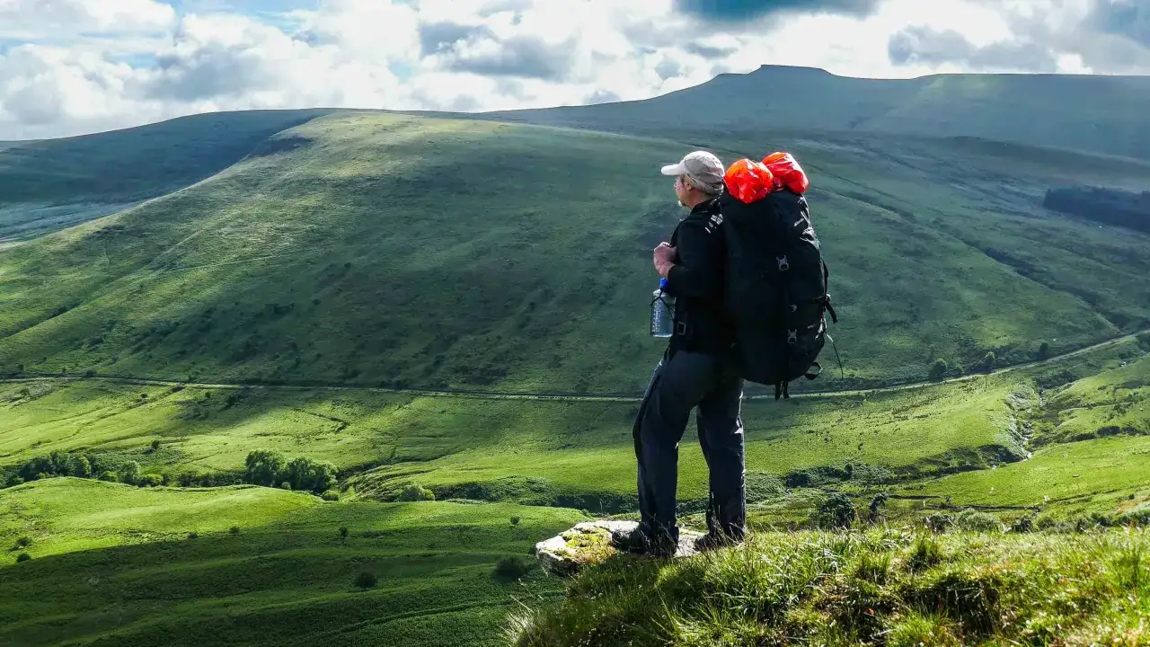 A hiker with a large backpack stands on a cliff edge, contemplating how to go backpacking through the vast, green, rolling hills under a cloudy sky.