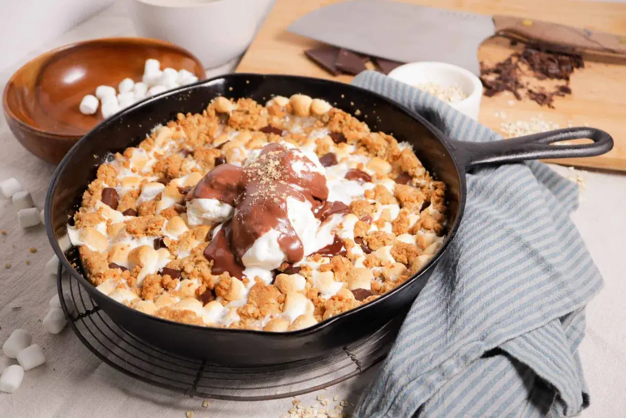 A warm smores pan dessert with melted marshmallows, chocolate chunks, and a scoop of ice cream drizzled with chocolate sauce.