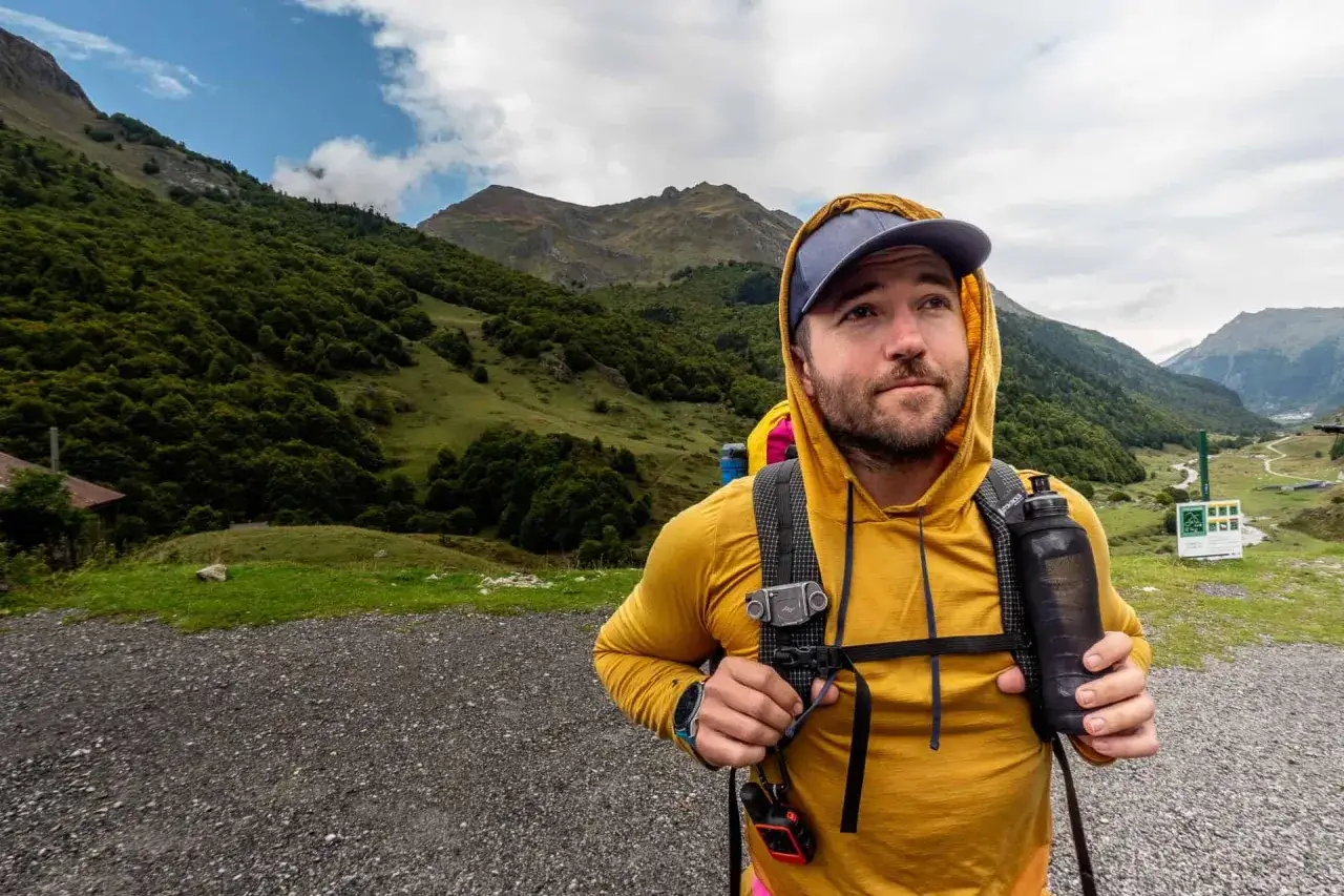 A hiker in a yellow hoodie, ready for adventure, demonstrates how to pack light with a minimalist backpack and water bottle, against a backdrop of lush green mountains.