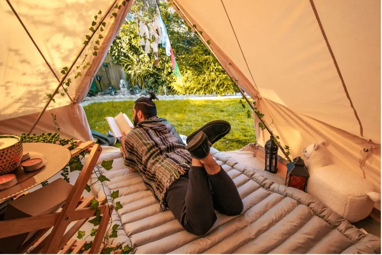 Man reading in a tent, with a cozy tent camping list setup including a mat, lanterns, and a plush sheep.