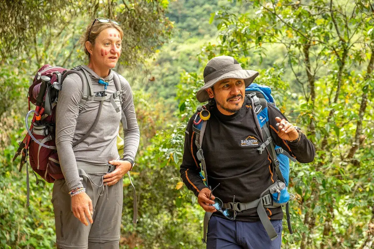 Hikers with backpacks, one with face paint, are carrying water on a trail through lush green foliage.