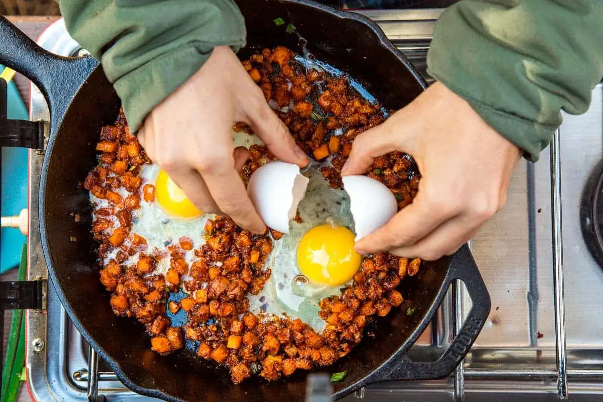 Cracking eggs into a cast iron skillet with diced sweet potatoes and spices. Delicious camping breakfast ideas in the making!