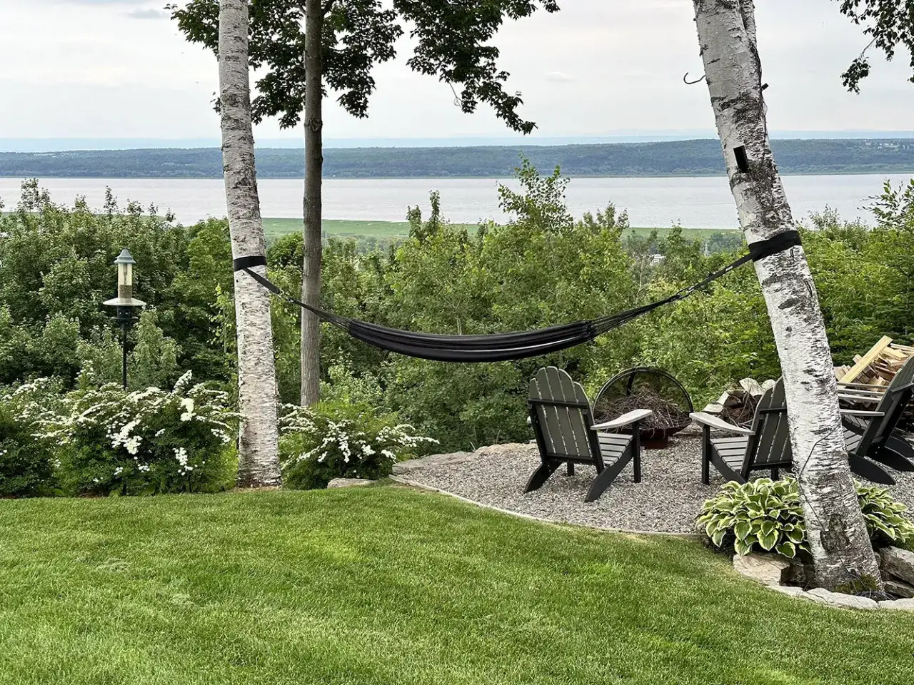 A black hammock is strung between two birch trees, demonstrating how to hang a hammock for a relaxing outdoor space with chairs and a fire pit overlooking a scenic water view.