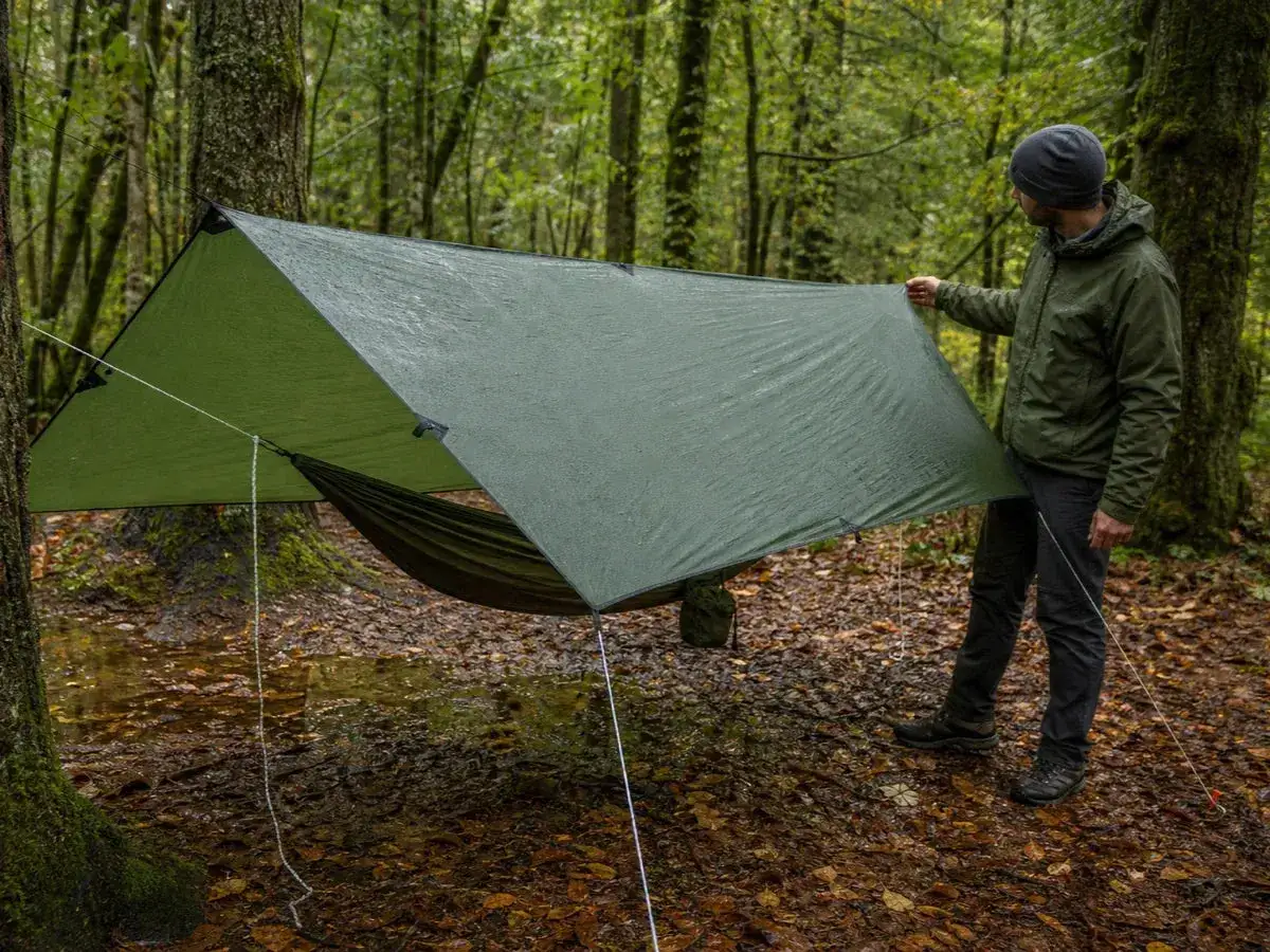 A man sets up a hammock camping setup in a wet forest. A green tarp is stretched over a hammock, providing shelter.