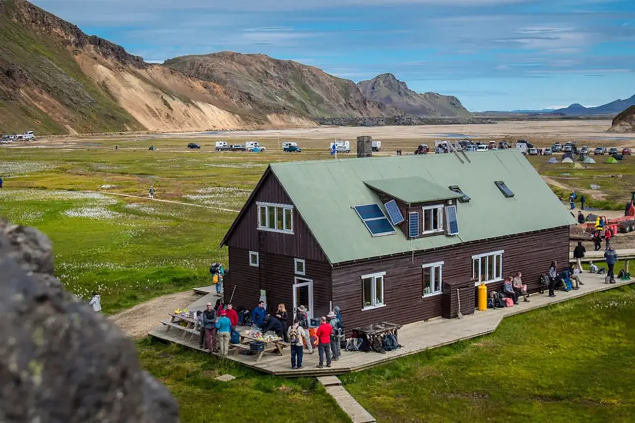 Hikers gather at a rustic hut along the Laugavegur trail in Iceland, with mountains and a vast landscape in the background.