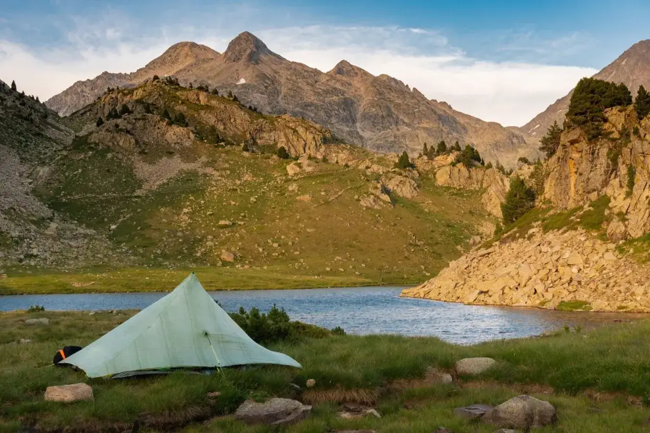 A lone tent sits by a serene lake, a perfect campsite on the Pyrenees High Route. Jagged peaks rise majestically in the background under a soft sky.