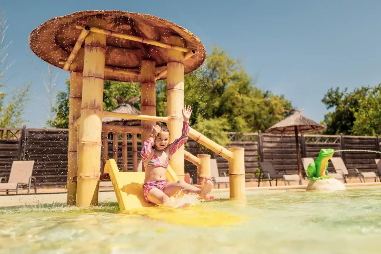 A girl gleefully slides down a yellow water slide at a family camping resort, splashing into the pool.