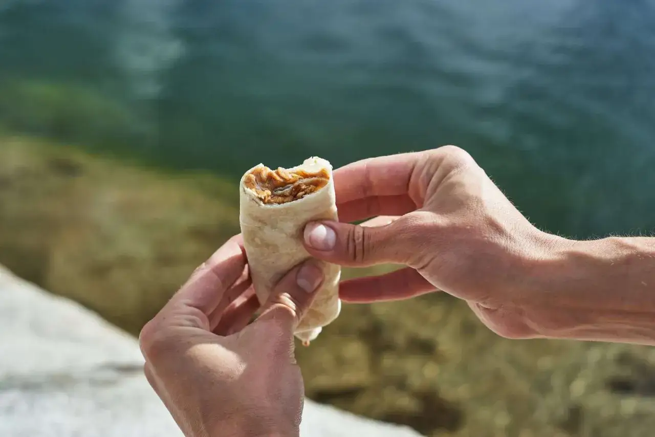 Hands hold a peanut butter and jelly wrap, a perfect no-cook backpacking lunch idea, by a serene body of water.