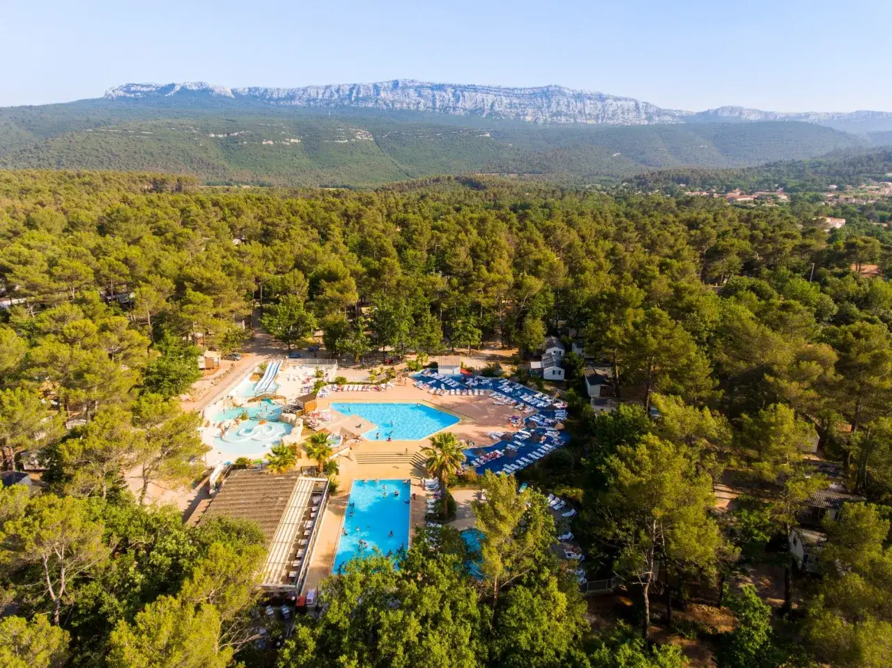 Aerial view of a resort with pools and water slides, nestled among trees with the Sainte Baume mountains in the background.