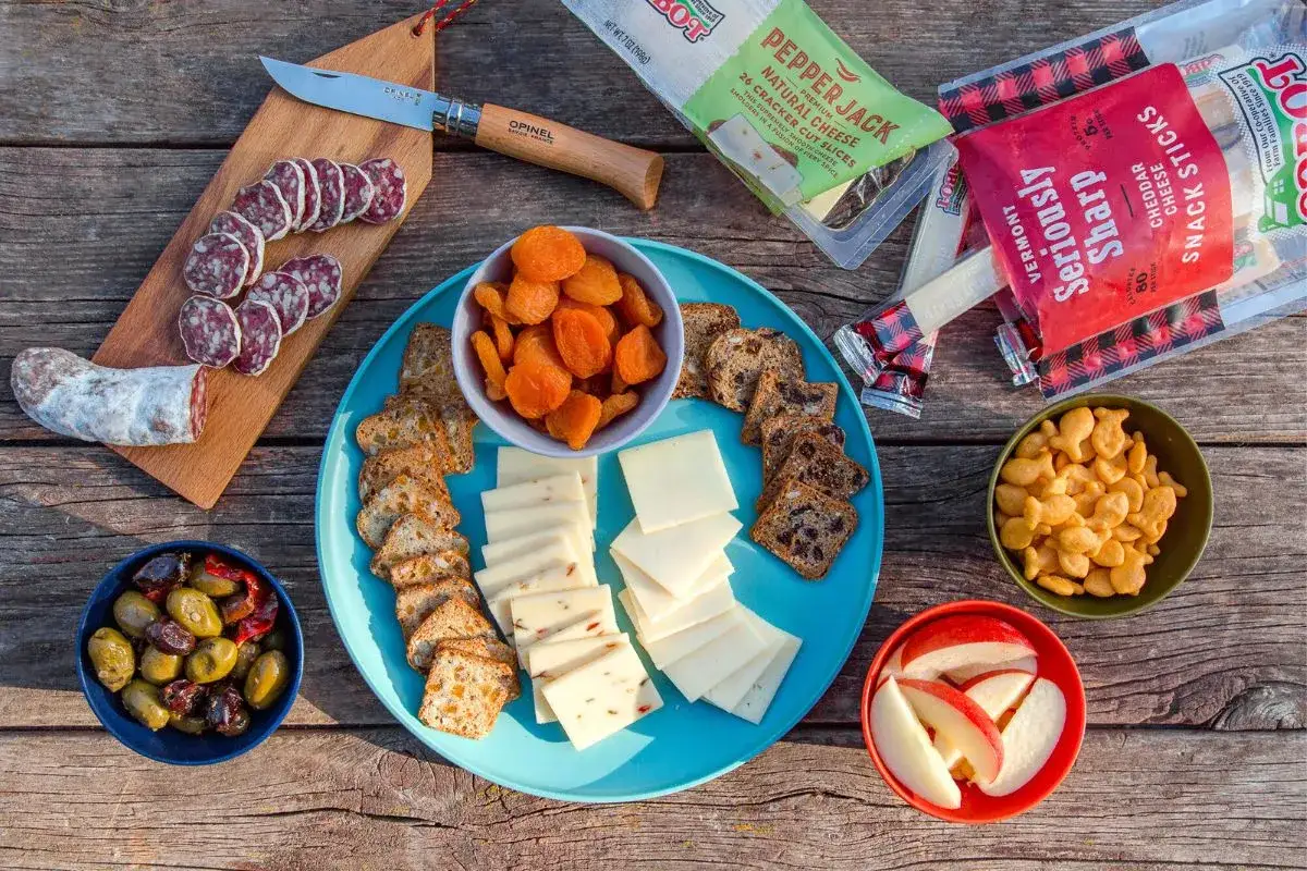 A delicious spread of camping snacks: sliced salami, cheese, crackers, dried apricots, olives, apple slices, and goldfish crackers.