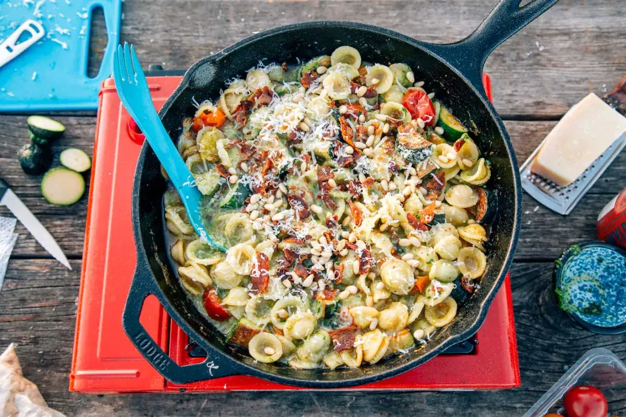 A delicious camping pasta dish with orecchiette, zucchini, tomatoes, bacon, pine nuts, and parmesan, served in a cast-iron skillet on a portable stove.