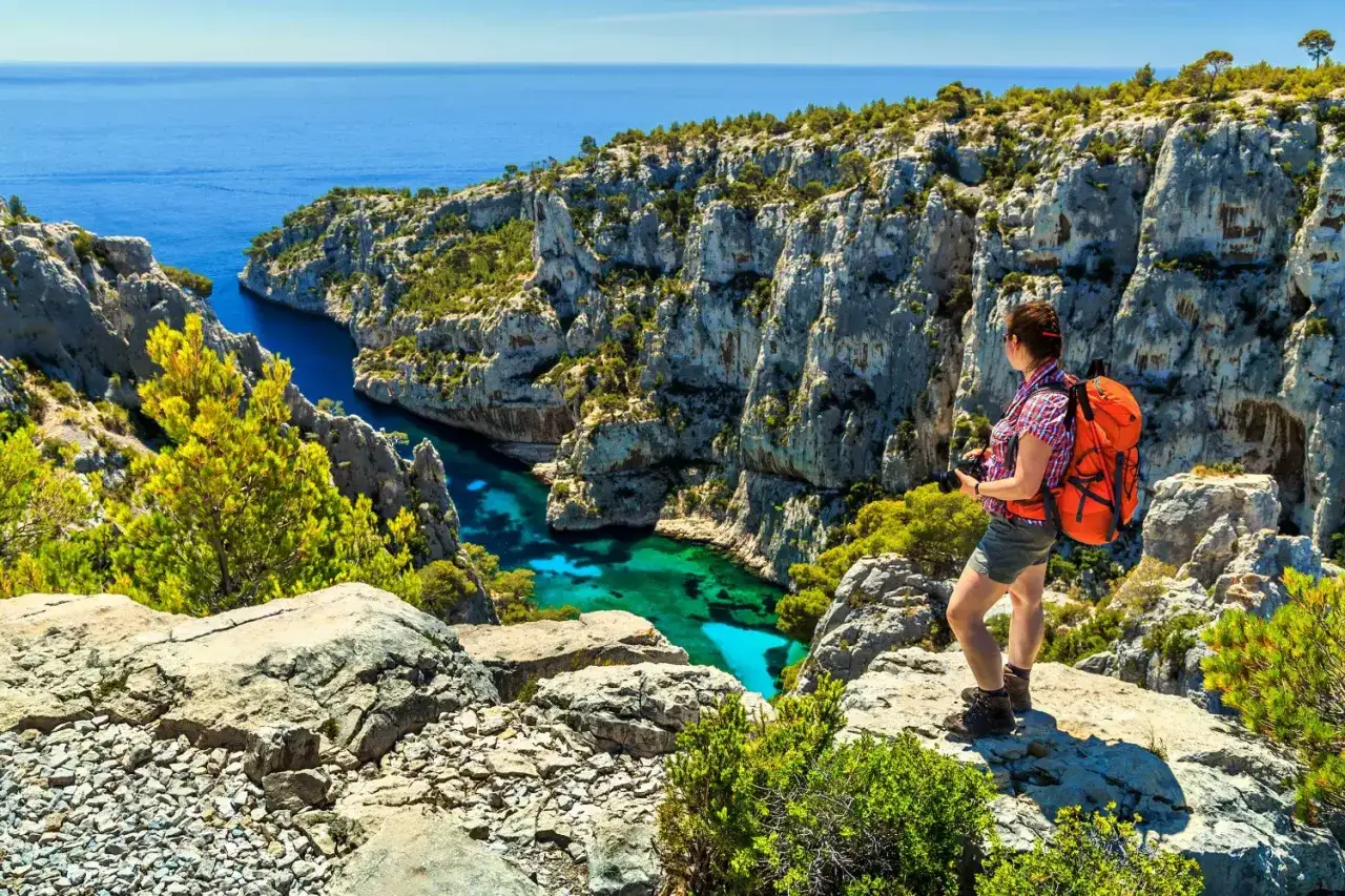 A hiker with an orange backpack pauses to photograph the stunning calanques of Cassis, with turquoise waters nestled between dramatic limestone cliffs.
