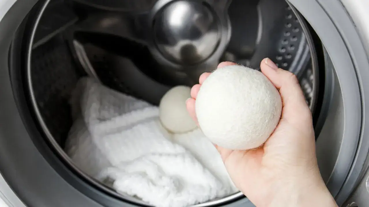 A hand holds a wool dryer ball, ready to add it to a washing machine with a white garment, part of learning how to wash a down jacket.