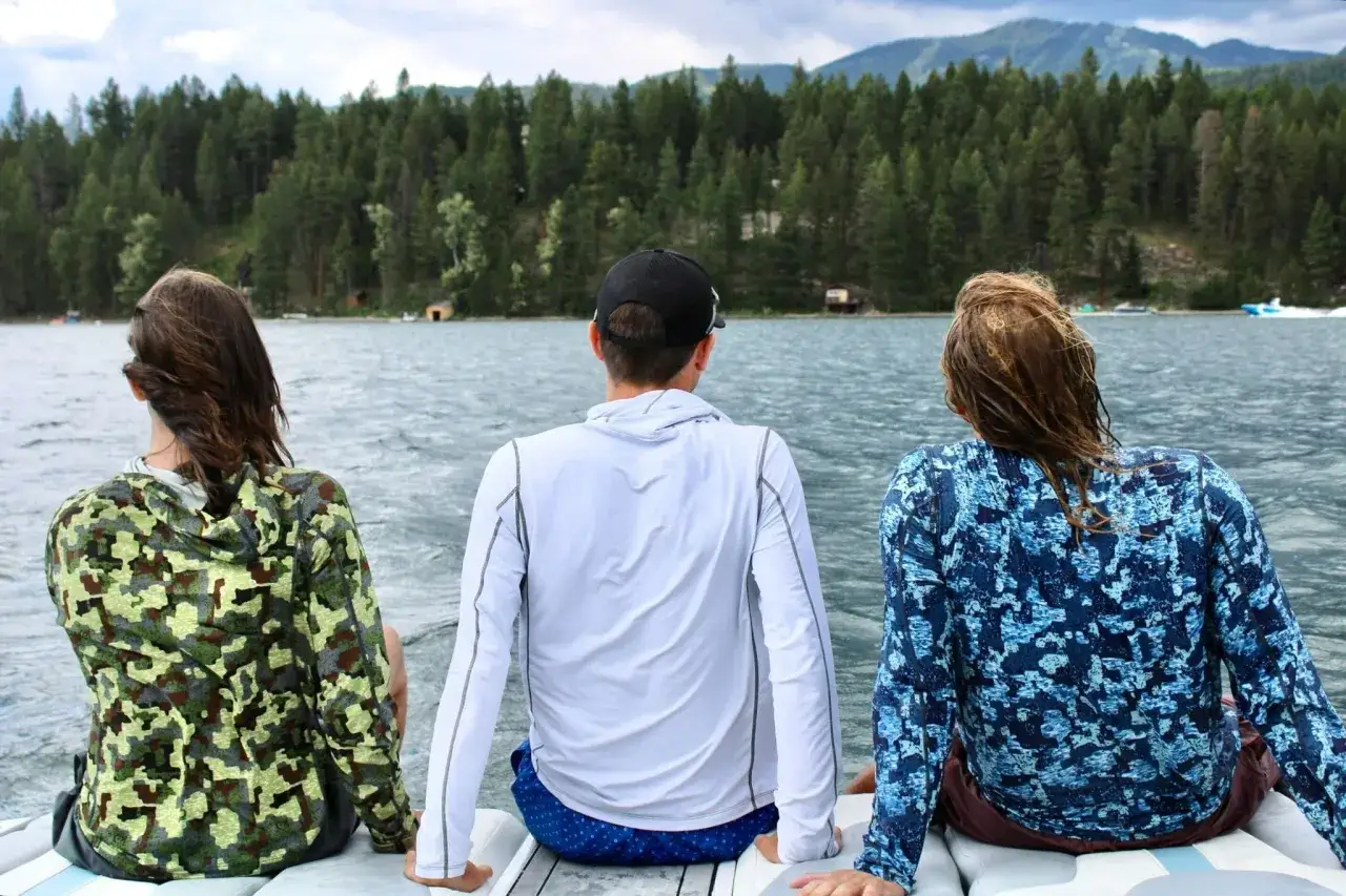 Three friends enjoy a lake view, wearing cooling clothing for extreme heat. The person on the left wears a camouflage hoodie, the middle wears a white long-sleeve shirt, and the right wears a blue patterned shirt.