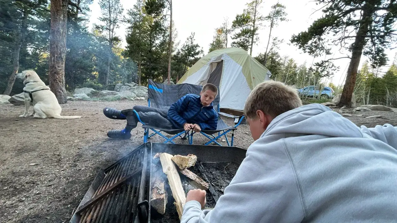 Boys relax by a campfire while a dog watches. The large tent in the background suggests that core tents are good quality for family camping trips.