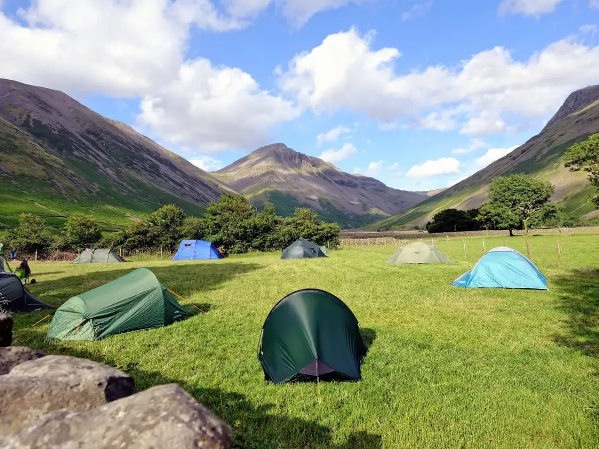 A large group camping setup in a grassy valley, with multiple tents pitched under a bright blue sky with fluffy clouds.