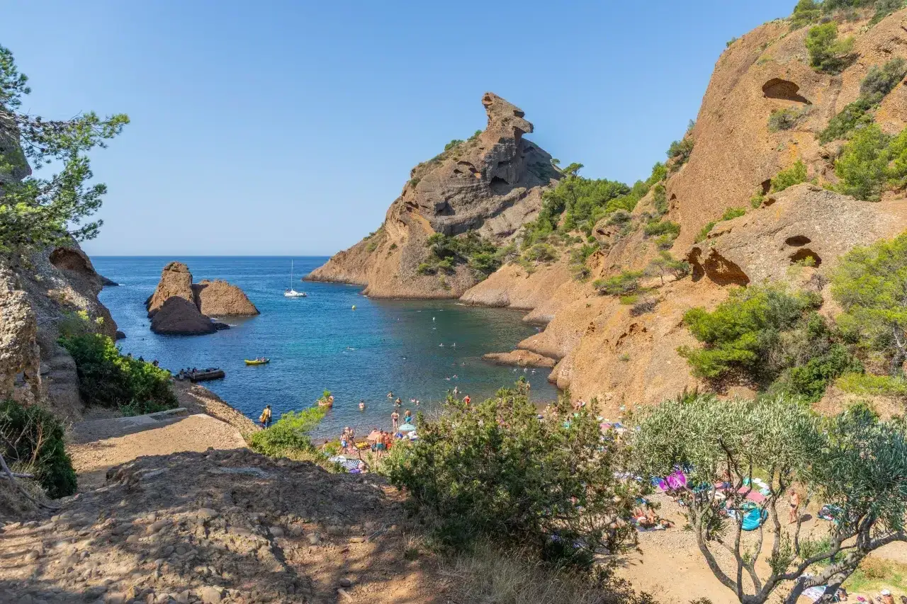 A beautiful bay near Cassis, with people enjoying a sunny day on the beach and in the clear blue water. The dramatic cliffs and rocky outcrops create a stunning backdrop for this idyllic scene.