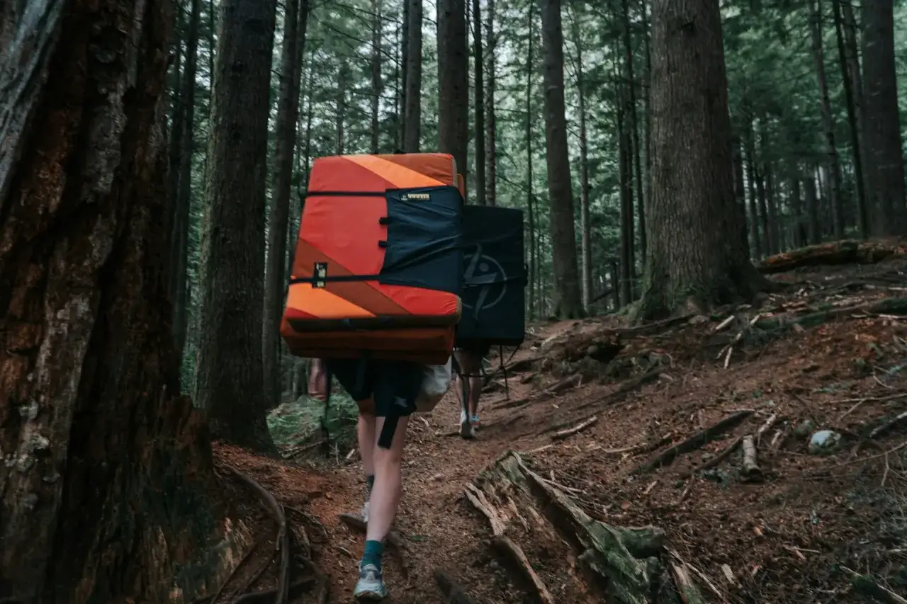 Hikers carry crash pads through a forest, preparing for a day of bouldering rock climbing.