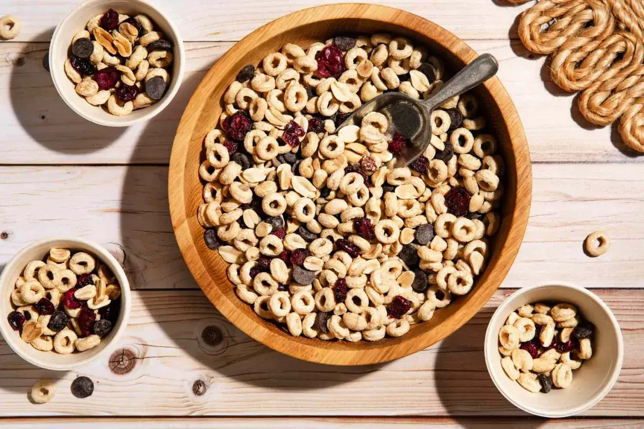 A wooden bowl and small bowls overflow with trail mix bar ingredients: O-shaped cereal, peanuts, dried cranberries, and chocolate chips.