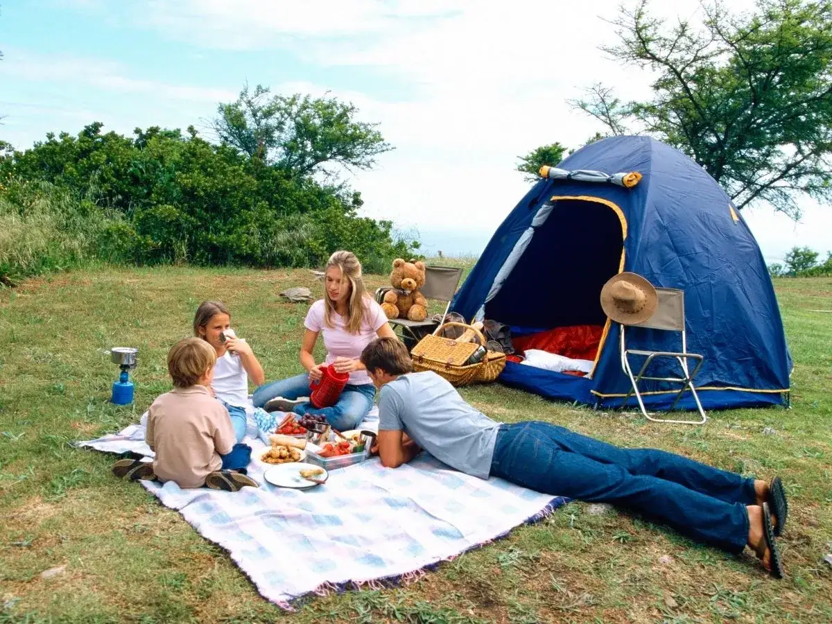 A family enjoys a picnic during camping with kids. A blue tent stands open, with a teddy bear and hat nearby.