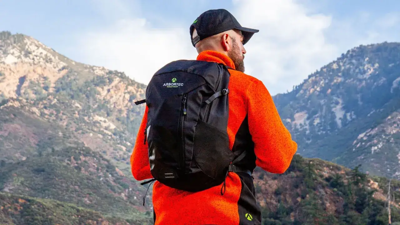 Man with a black backpack and orange jacket looks out at mountains, ready for beginner backpacking.