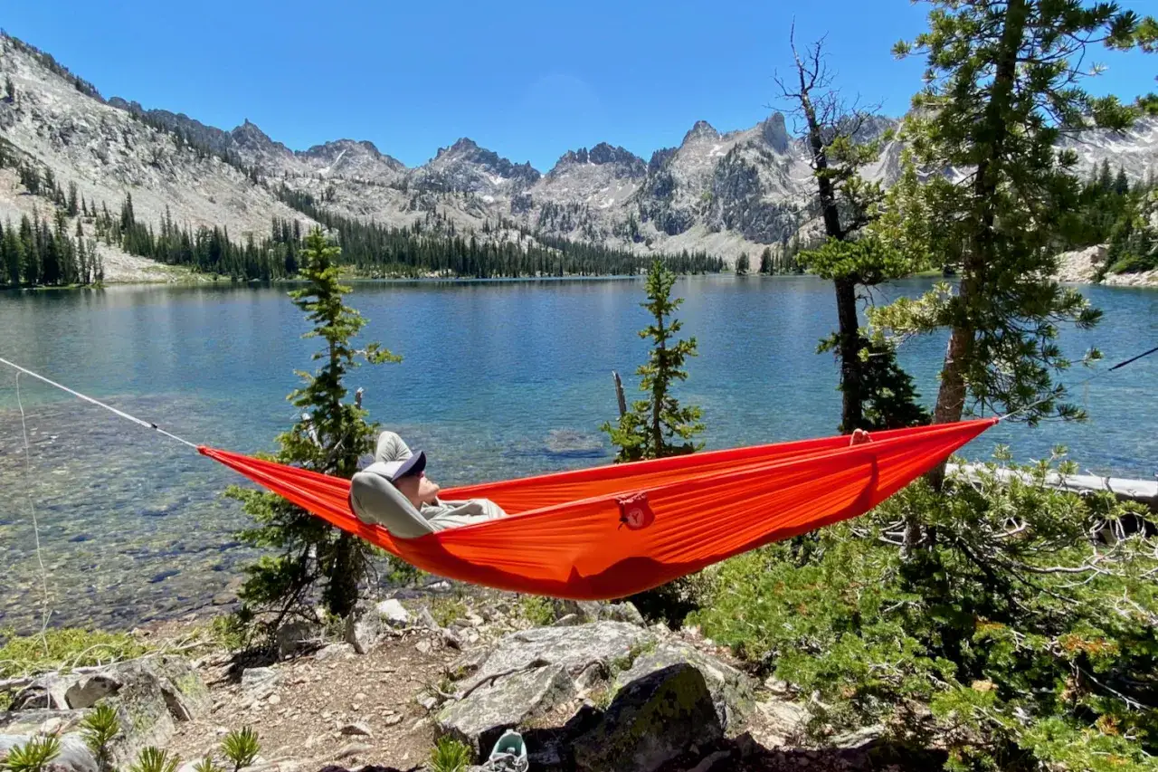 A person relaxes in a bright orange double hammock by a clear blue lake, with a majestic mountain range in the background.