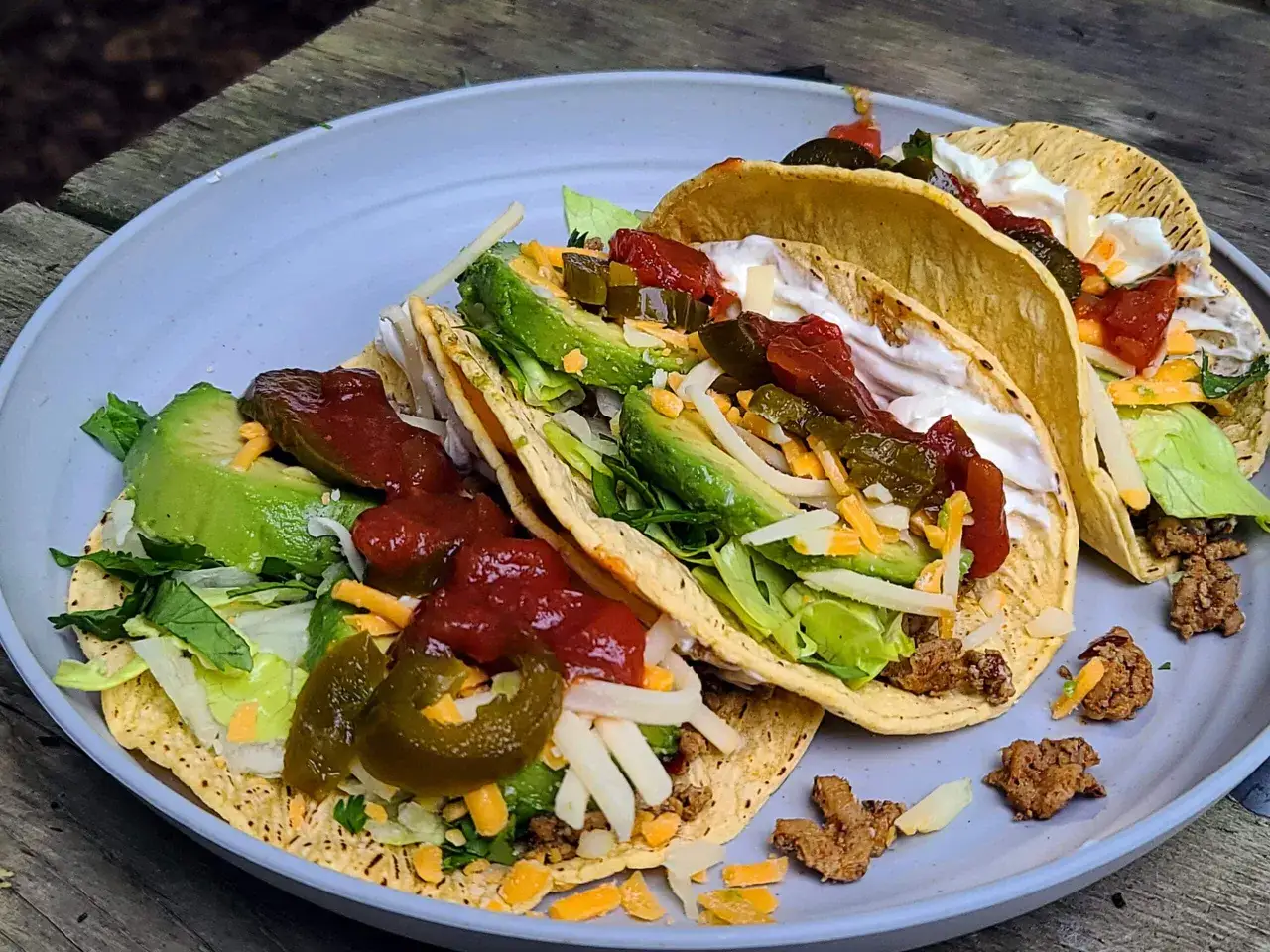 Delicious camping tacos piled high with ground beef, lettuce, avocado, cheese, salsa, and jalape&ntilde;os on a rustic wooden table.